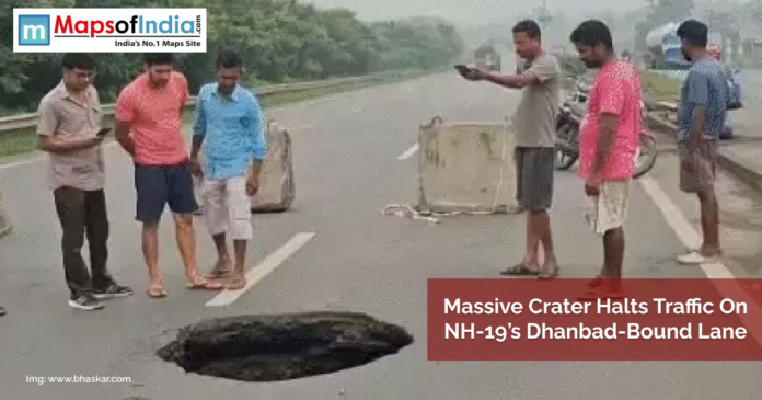 People gather around a large crater that has formed on NH-19's Dhanbad-bound lane, causing a traffic halt; concrete barriers are placed nearby for safety.