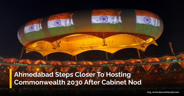 A large stadium illuminated at night with a giant overhead structure decorated with multiple Indian flags. The stadium is filled with spectators. Text overlay reads, 