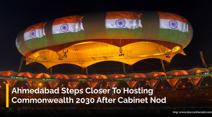 Ahmedabad Steps Closer to Hosting Commonwealth 2030 After Cabinet Nod A large stadium illuminated at night with a giant overhead structure decorated with multiple Indian flags. The stadium is filled with spectators. Text overlay reads, "Ahmedabad Steps Closer To Hosting Commonwealth 2030 After Cabinet Nod."