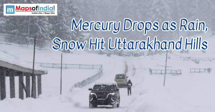 Mercury drops in Uttarakhand Snow-covered road in the hills of Uttarakhand with vehicles and people navigating through heavy snowfall. The headline reads, “Mercury Drops as Rain, Snow Hit Uttarakhand Hills,” with the Maps of India logo in the corner.