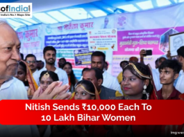 A senior official interacts with a group of women dressed in traditional attire at a public event, with banners displayed in the background.
