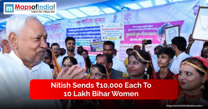 A senior official interacts with a group of women dressed in traditional attire at a public event, with banners displayed in the background.
