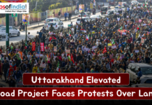 Uttarakhand Elevated Road Project Faces Protests Over Land Large crowd of protesters marching along a busy Uttarakhand street, holding placards and flags as traffic lines the road, highlighting opposition to the elevated road project over land acquisition.