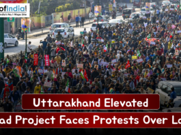 Large crowd of protesters marching along a busy Uttarakhand street, holding placards and flags as traffic lines the road, highlighting opposition to the elevated road project over land acquisition.