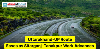 Uttarakhand-UP Route Eases as Sitarganj–Tanakpur Work Advances A scenic multi-lane highway curves through a lush, green hilly landscape under overcast skies. The image includes the Maps of India logo in the top left and a yellow headline reading "Uttarakhand-UP Route Eases as Sitarganj–Tanakpur Work Advances."