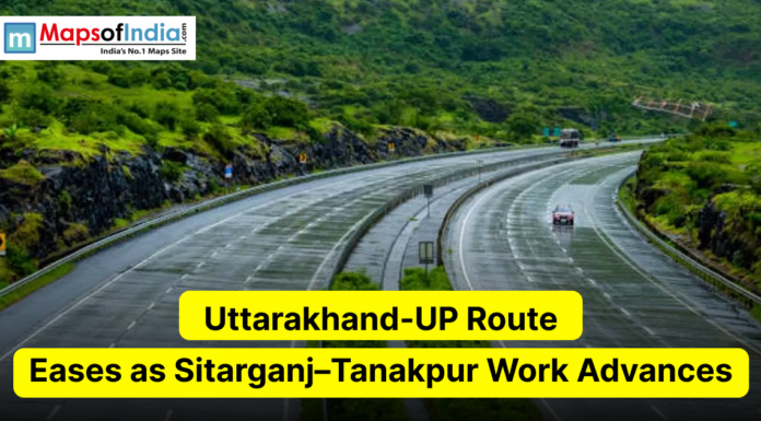 Uttarakhand-UP Route Eases as Sitarganj–Tanakpur Work Advances A scenic multi-lane highway curves through a lush, green hilly landscape under overcast skies. The image includes the Maps of India logo in the top left and a yellow headline reading "Uttarakhand-UP Route Eases as Sitarganj–Tanakpur Work Advances."