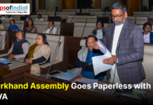 Jharkhand Assembly Goes Paperless with NeVA Jharkhand Chief Minister speaking in the state assembly hall while holding documents, with legislators seated at desks wearing headphones; banner reads “Jharkhand Assembly Goes Paperless with NeVA” and includes the Maps of India logo.