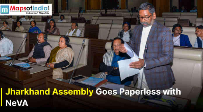 Jharkhand Assembly Goes Paperless with NeVA Jharkhand Chief Minister speaking in the state assembly hall while holding documents, with legislators seated at desks wearing headphones; banner reads “Jharkhand Assembly Goes Paperless with NeVA” and includes the Maps of India logo.
