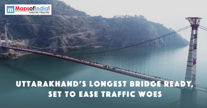 A long suspension bridge over a river in Uttarakhand, surrounded by hills, with text announcing it as the state’s longest bridge ready to ease traffic.